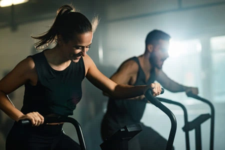 Femme sportive en plein effort, pratiquant le vélo Air Bike avec un autre sportif en arrière plan.